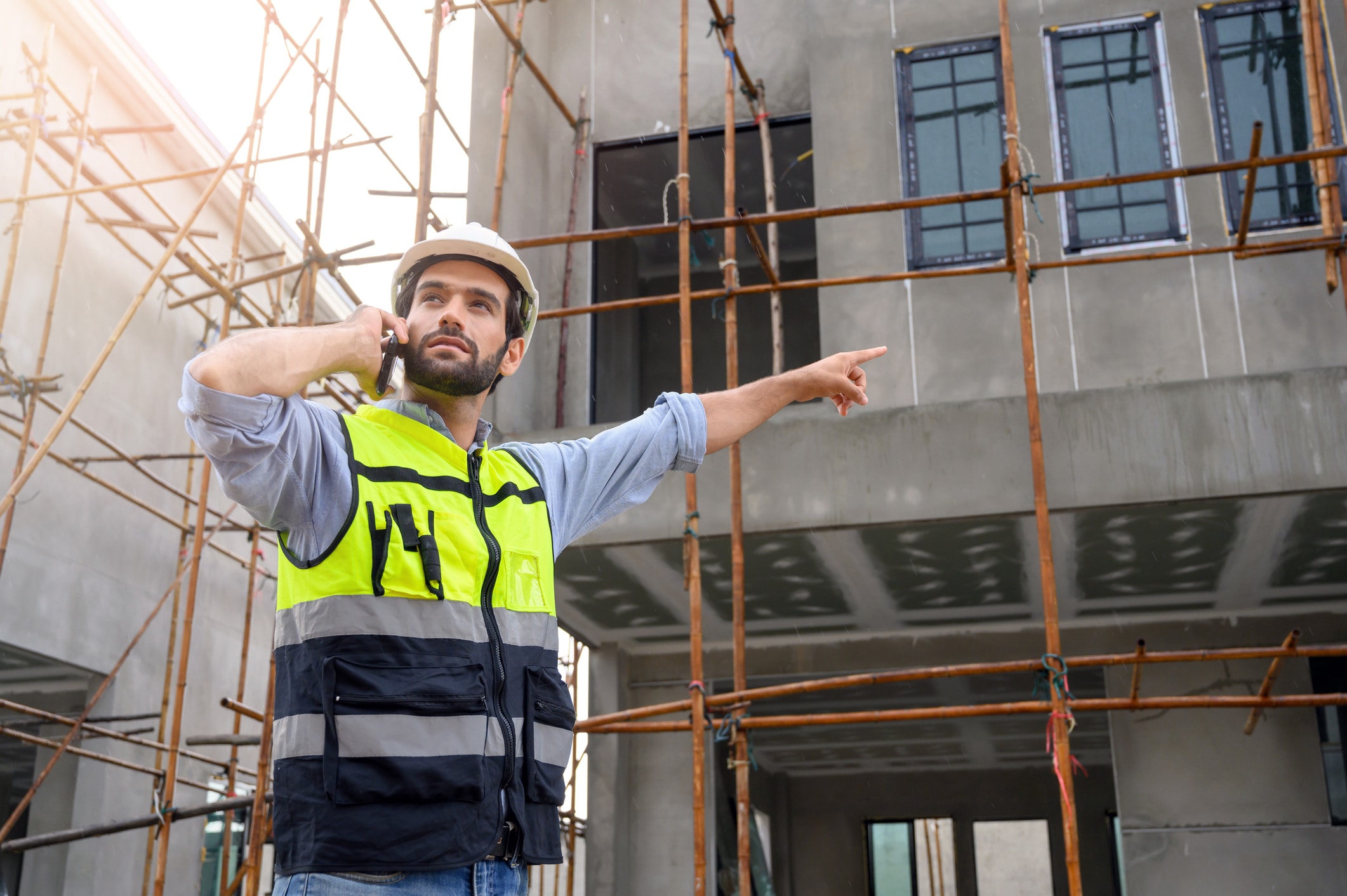 Young attractive construction smiling bearded using smartphone calling to client, worker in vest with white helmet standing on construction site.