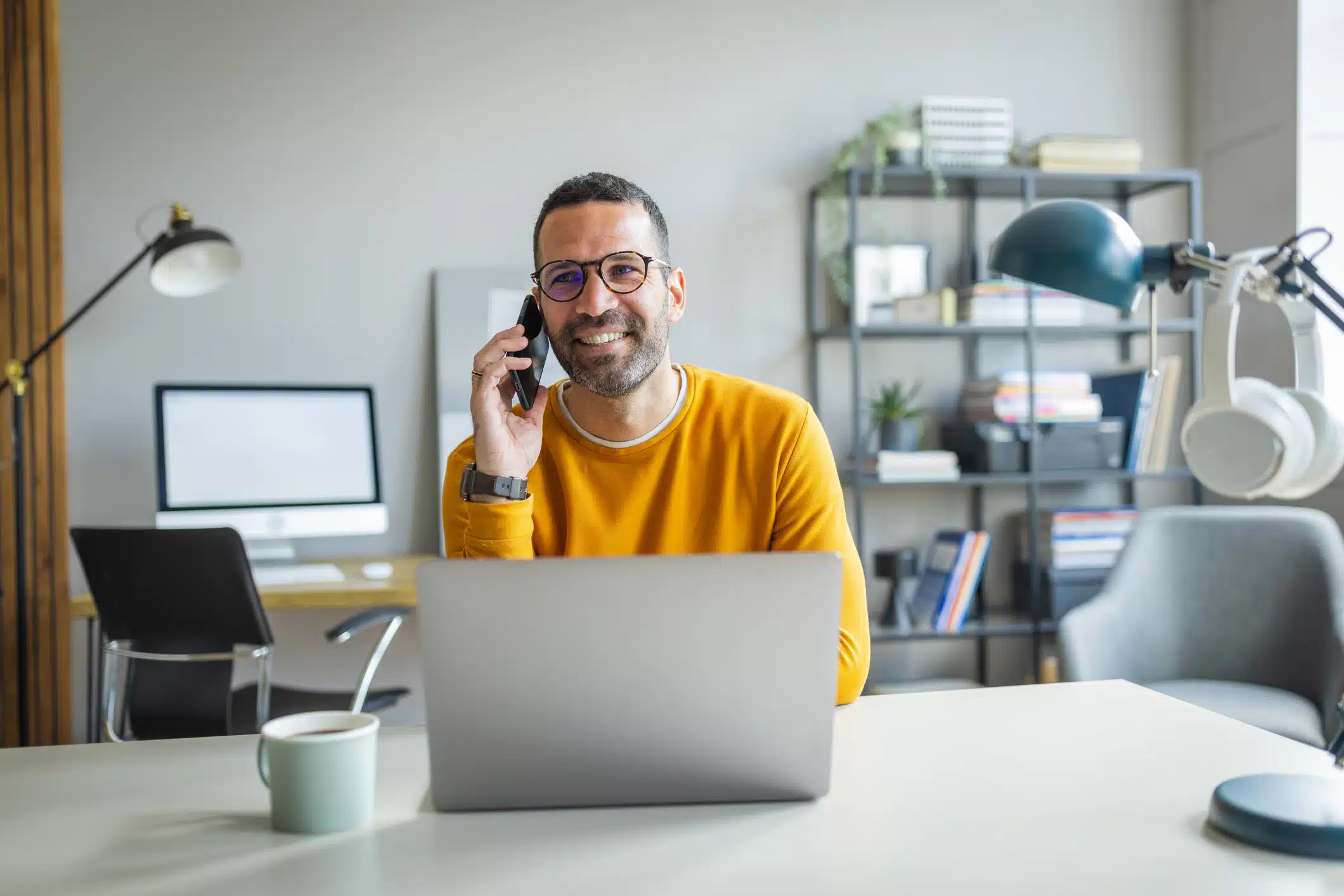Empresario con ropa informal y gafas sentado en su escritorio, usando un portátil y un teléfono inteligente en la oficina.